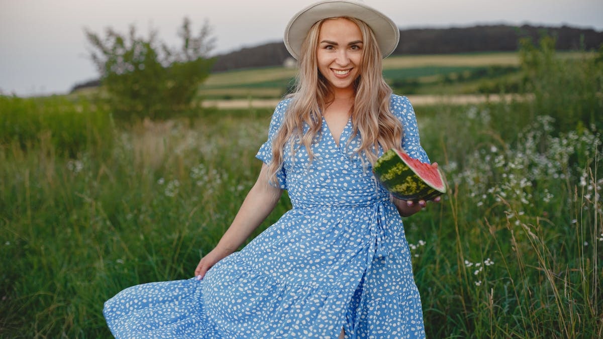 Happy woman in a summer dress and hat holding watermelon in a scenic outdoor meadow.
